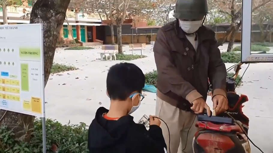 Một bạn học sinh nhờ người lớn nổ xe máy để đo chất lượng không khí gần ống xả.